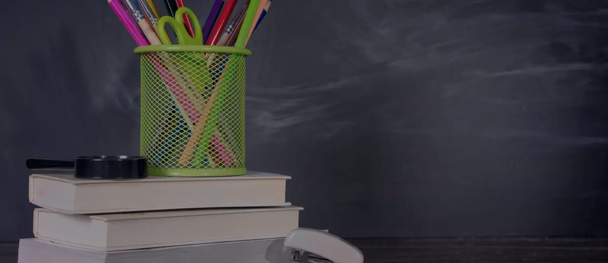 A stack of books with pencils and school supplies in front of a chalkboard.