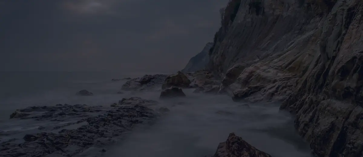 Rocky sea cliffs rising above misty waves under a dim evening sky.