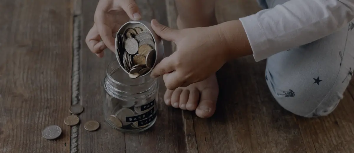 A child's hands dropping coins into a glass jar on a wooden floor.