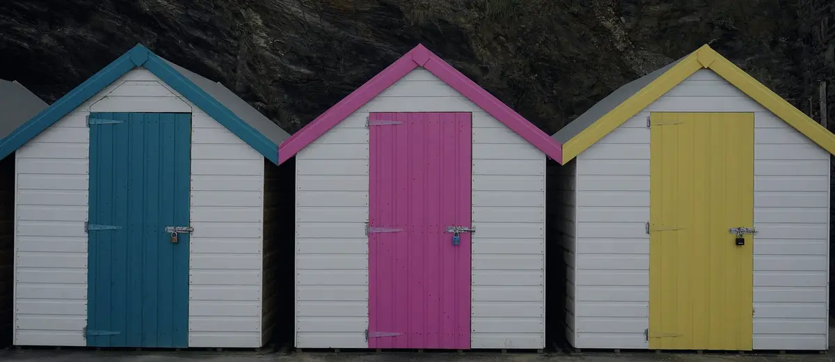 Three beach huts with blue, pink and yellow doors against dark rock.