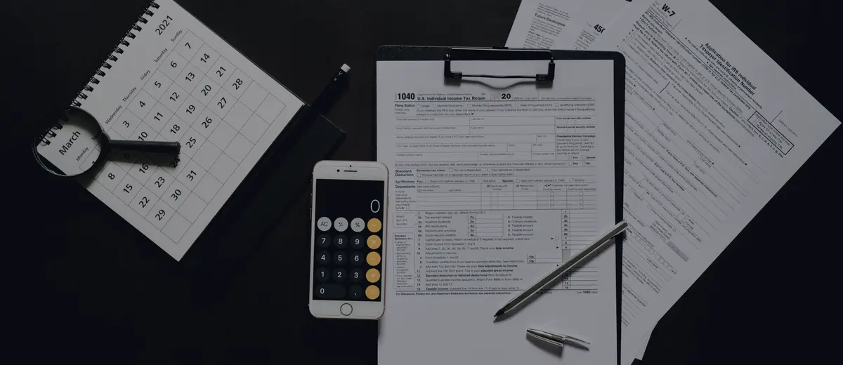 Tax forms on a clipboard beside a calculator, calendar and magnifying glass on a dark desk.