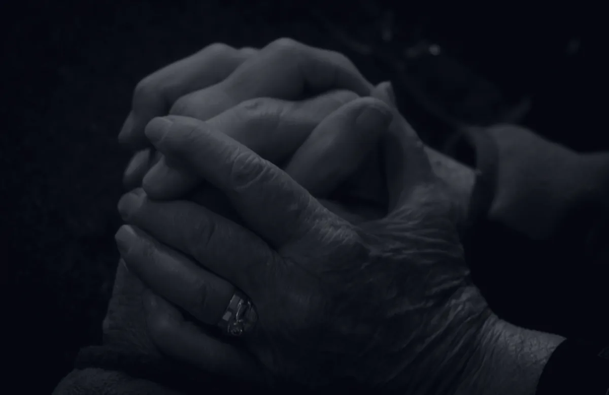 Clasped elderly and younger hands in dark tones, representing the finances of eldercare.