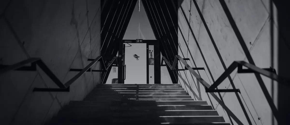 A black-and-white staircase climbing toward a bright doorway between angled concrete walls.