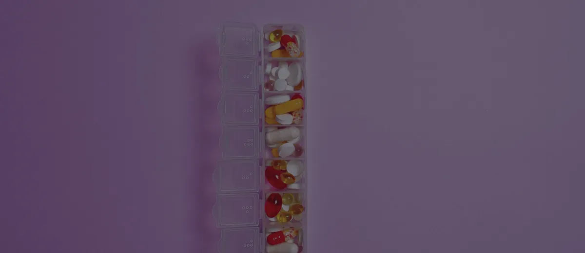 Assorted pills in a weekly organizer on a violet background: your prescription list drives the 2027 drug-plan choice.