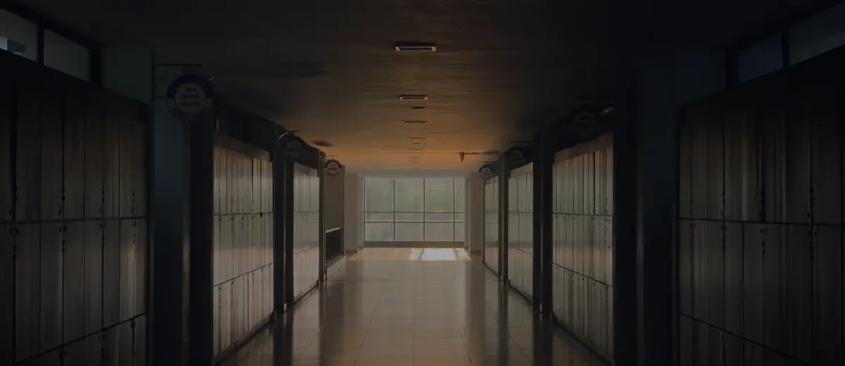 A dim school corridor lined with lockers, lit by late-afternoon sun at the far end.