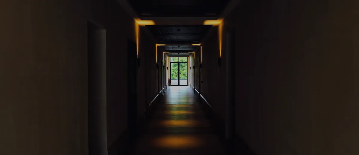 A dark hallway lit by a row of lights leading to a bright exit.