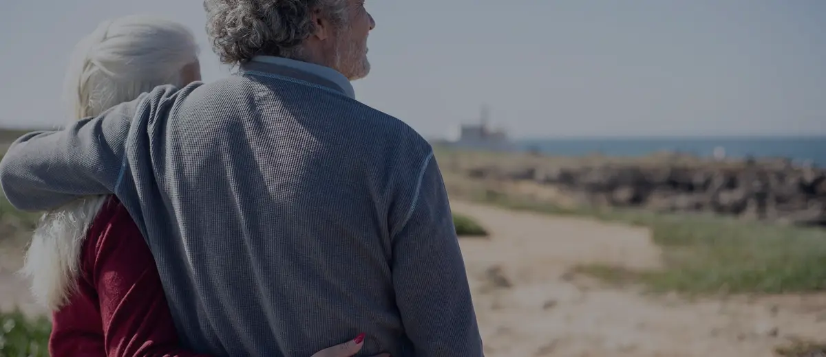 An older couple with their arms around each other, looking out at the sea.