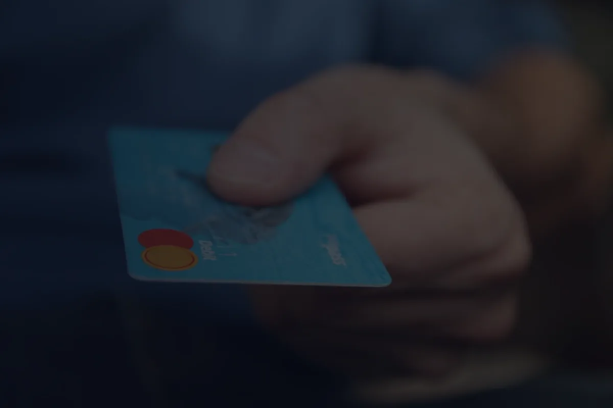 A hand holding a generic teal credit card against a dark background, illustrating credit card debt and minimum payments.