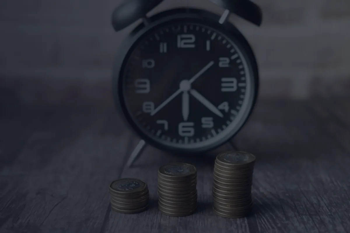 A dark alarm clock beside ascending stacks of coins on a wooden table, representing the timing trade-off of claiming Social Security.